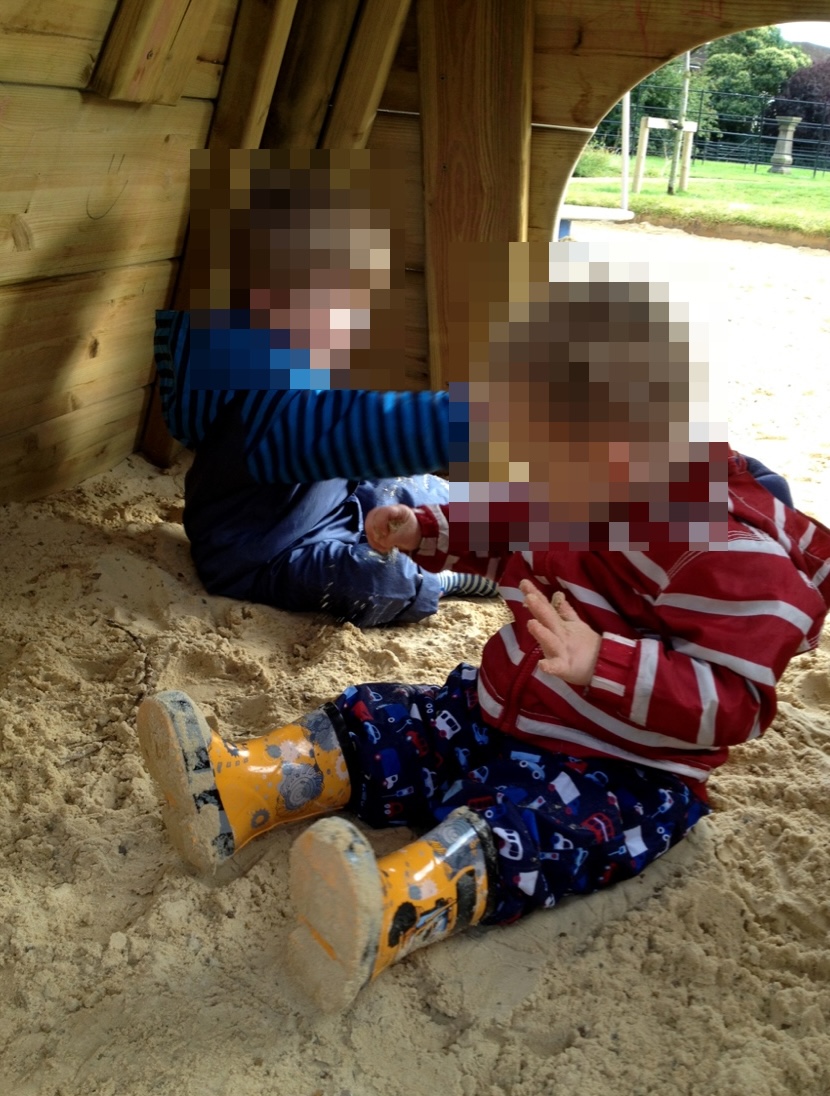 Two young children sitting in the sand inside a wooden play teepee structure, wearing coats and yellow wellington boots.