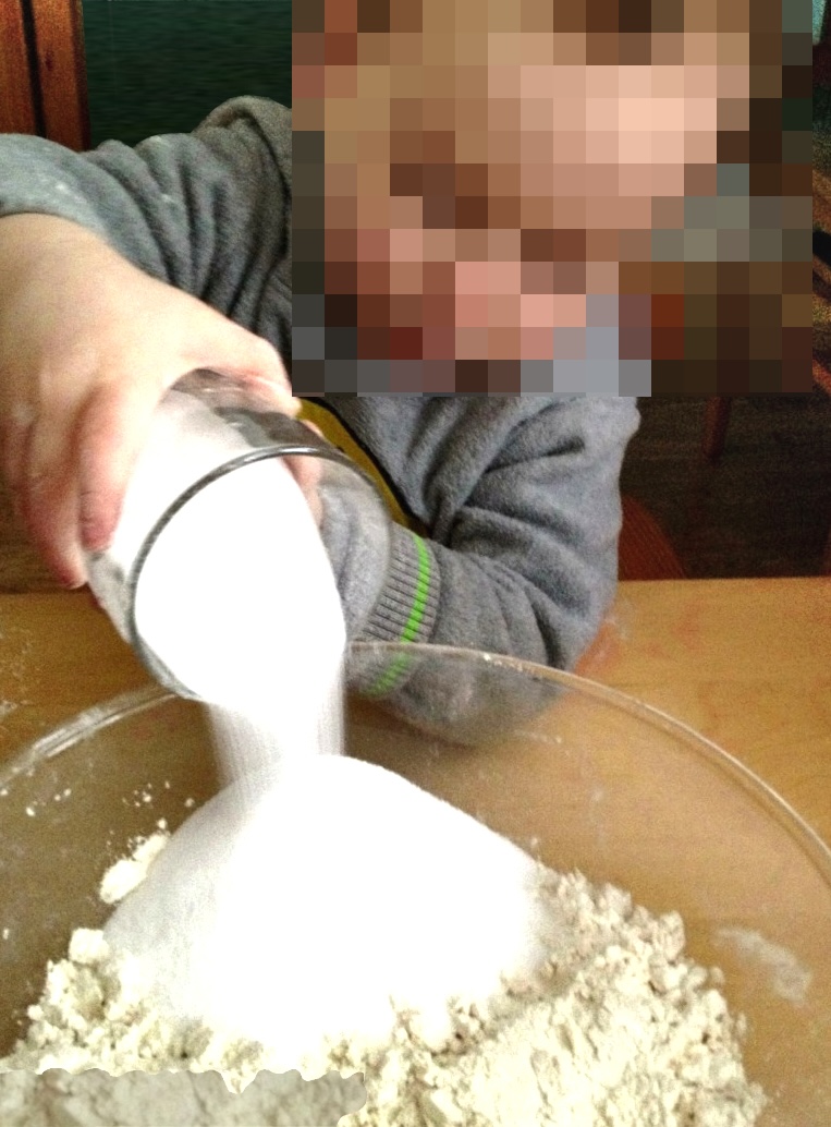 A young child wearing a grey sweater pouring white sugar or flour from a clear glass tumbler into a large mixing bowl.