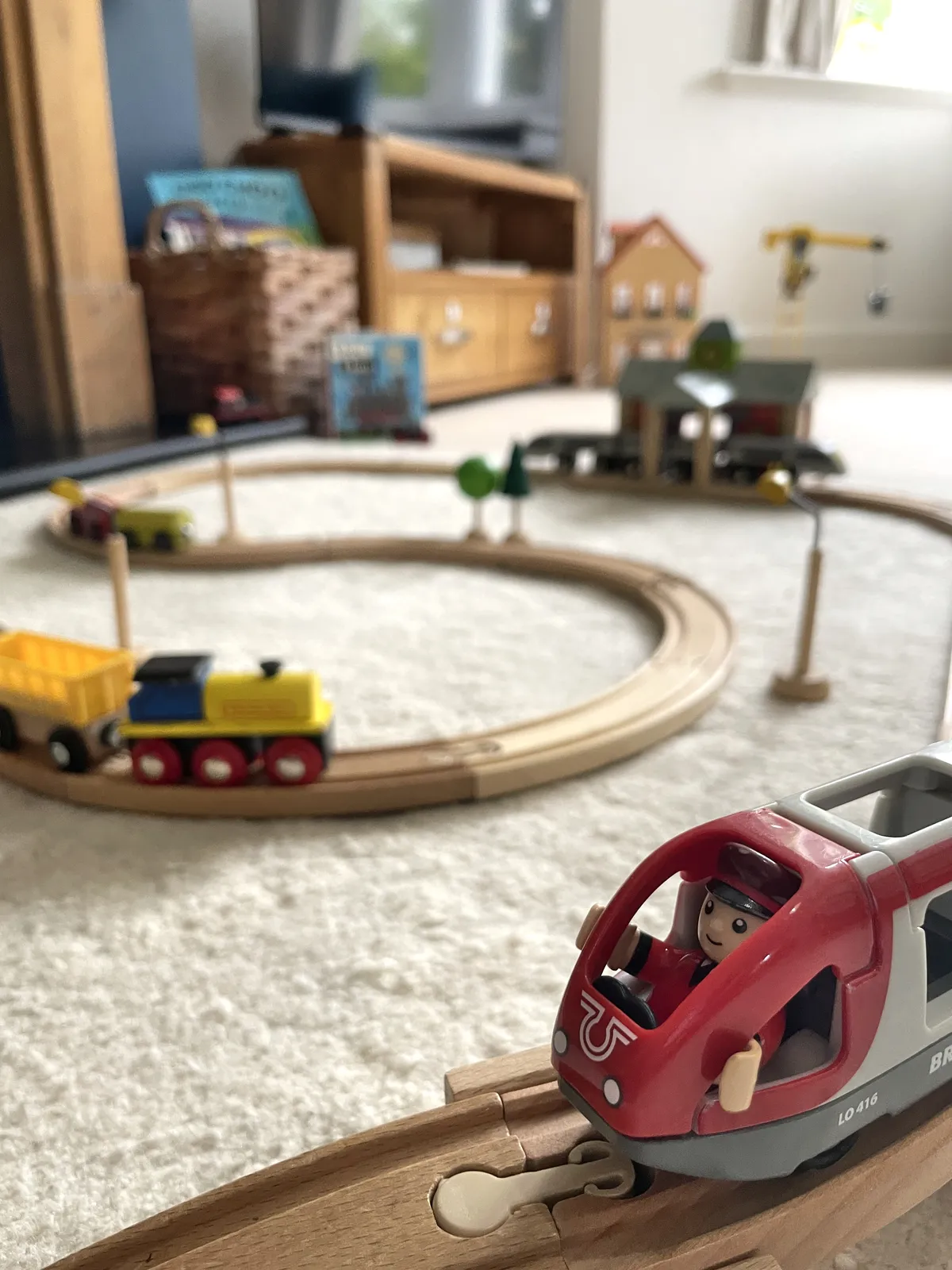 Brio toy train with driver in foreground driving train around wooden train track visible in background.