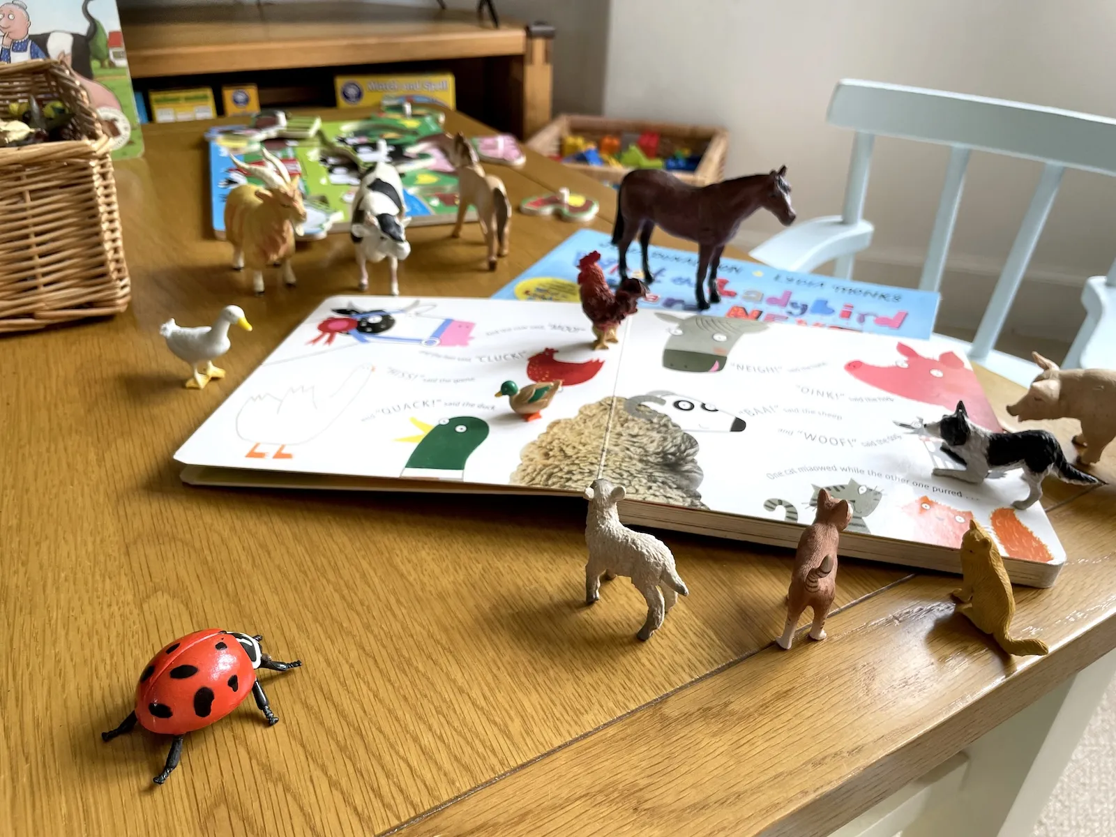 Farm themed toys on a wooden table top surrounding the What the ladybird heard story book and the characters from that story. A farm puzzle is further back and a children's chair visible.
