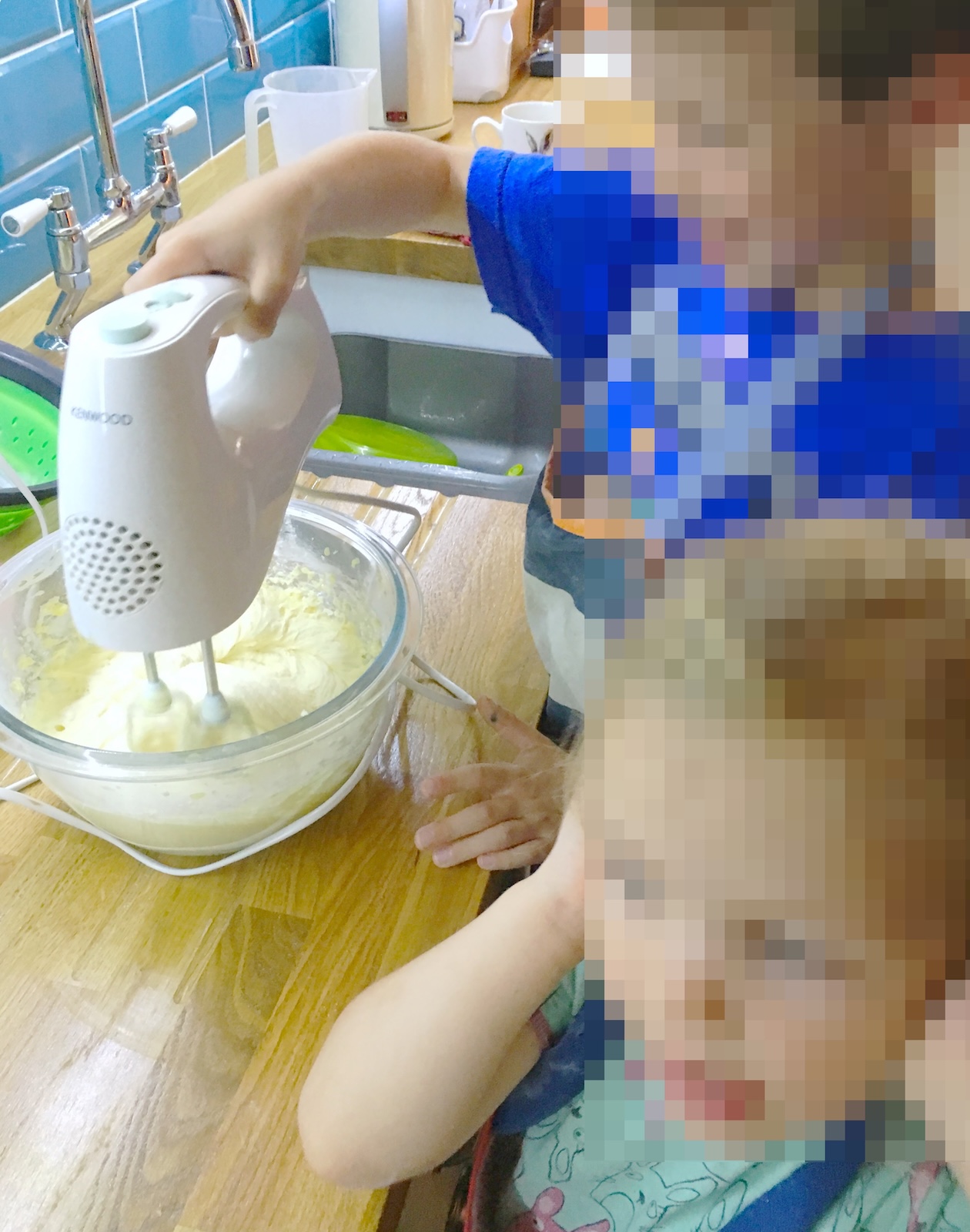 A young child uses a white electric hand mixer to blend batter in a glass bowl on a kitchen counter while another child covers their ears nearby.