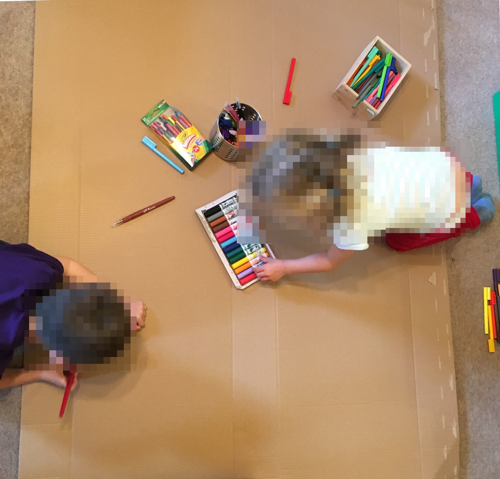 Overhead view of two young children lying on a large sheet of cardboard on the floor, surrounded by markers and crayons for drawing.
