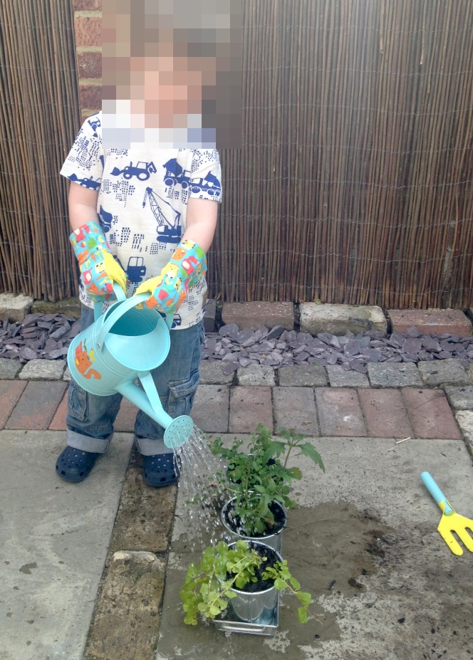 A young child wearing colourful gardening gloves uses a small light-blue watering can to water potted plants on a patio floor.