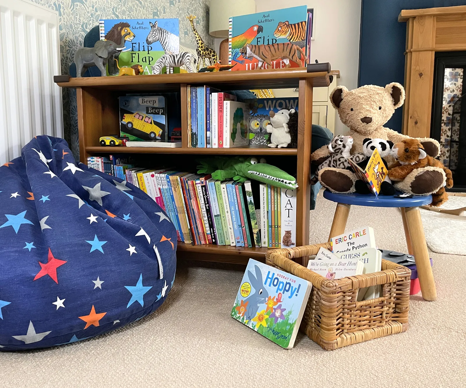 Reading shelf with star patterned bean bag, books and soft toys as well as animal flap books positioned on top with animal toys to match to play with. A teddy on a stool reading a Maisy Mouse book with his friends. A book basket on the carpet in front.