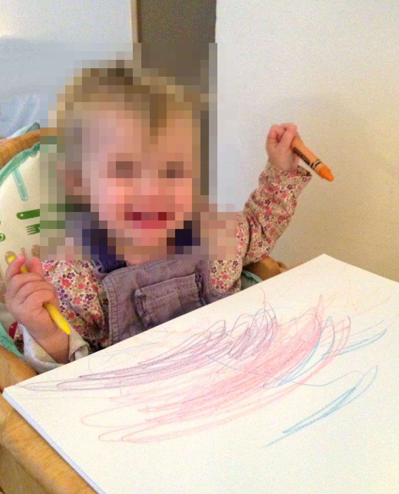 A young toddler sitting in a high chair holding yellow and orange crayons above a sheet of white paper filled with colourful scribbles.