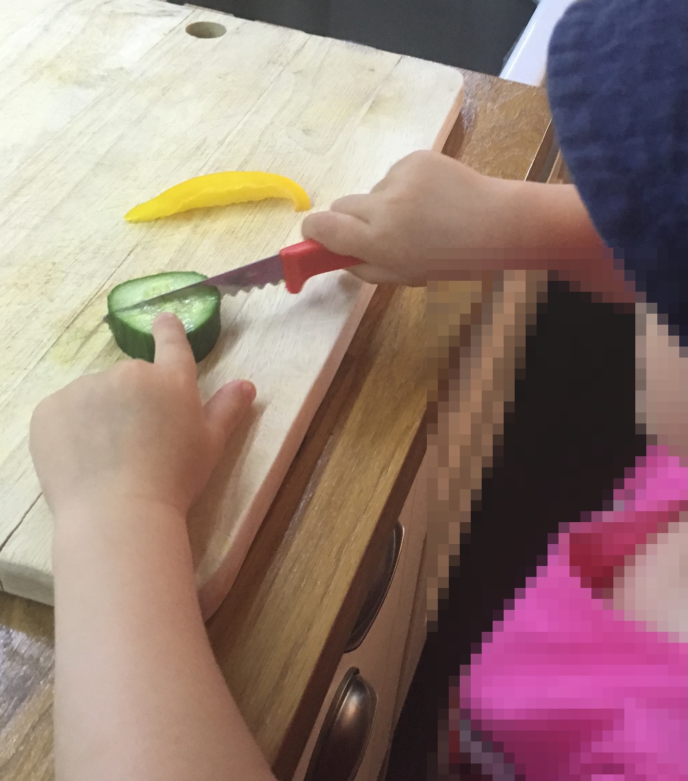 A young child wearing a pink top using a small red child-safe serrated knife to cut a cucumber slice on a wooden chopping board.