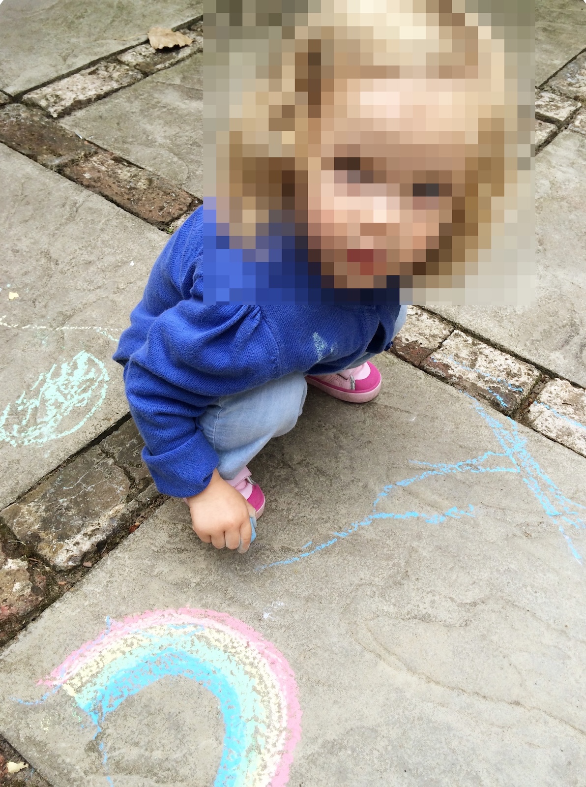 A young toddler crouching outdoors on stone paving slabs while drawing with chalk next to a chalk rainbow.