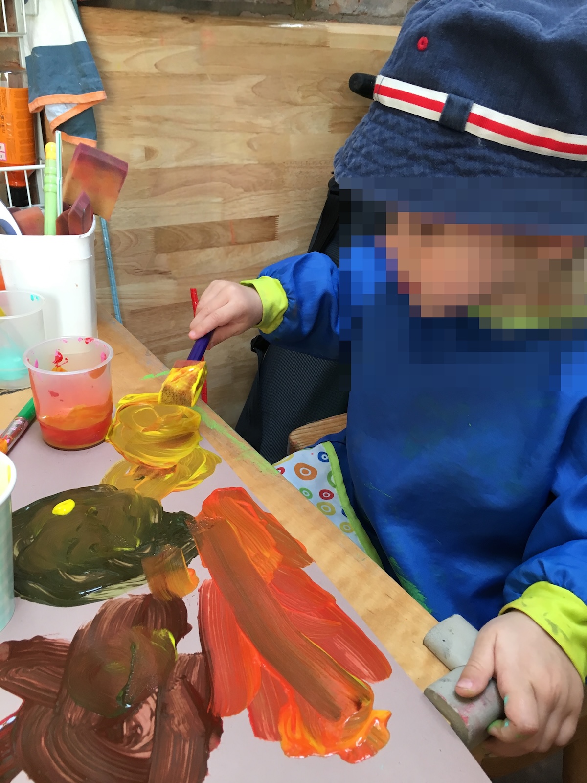 A young child wearing a blue smock and sun hat uses a foam sponge brush to spread yellow paint onto a paper covered in bright, thick acrylic paint swirls.