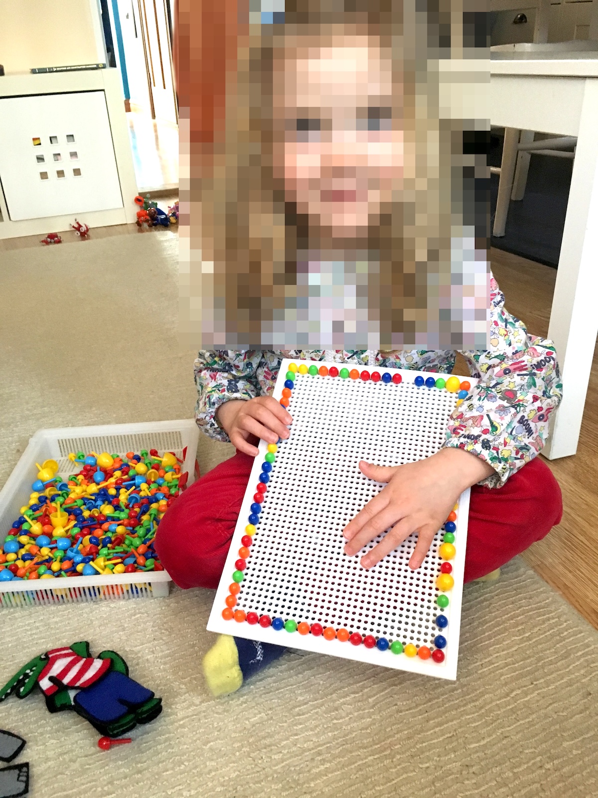 A young child sitting cross-legged on a rug holding a pegboard with a neat border of colorful plastic pegs, next to a bin full of extra pegs.