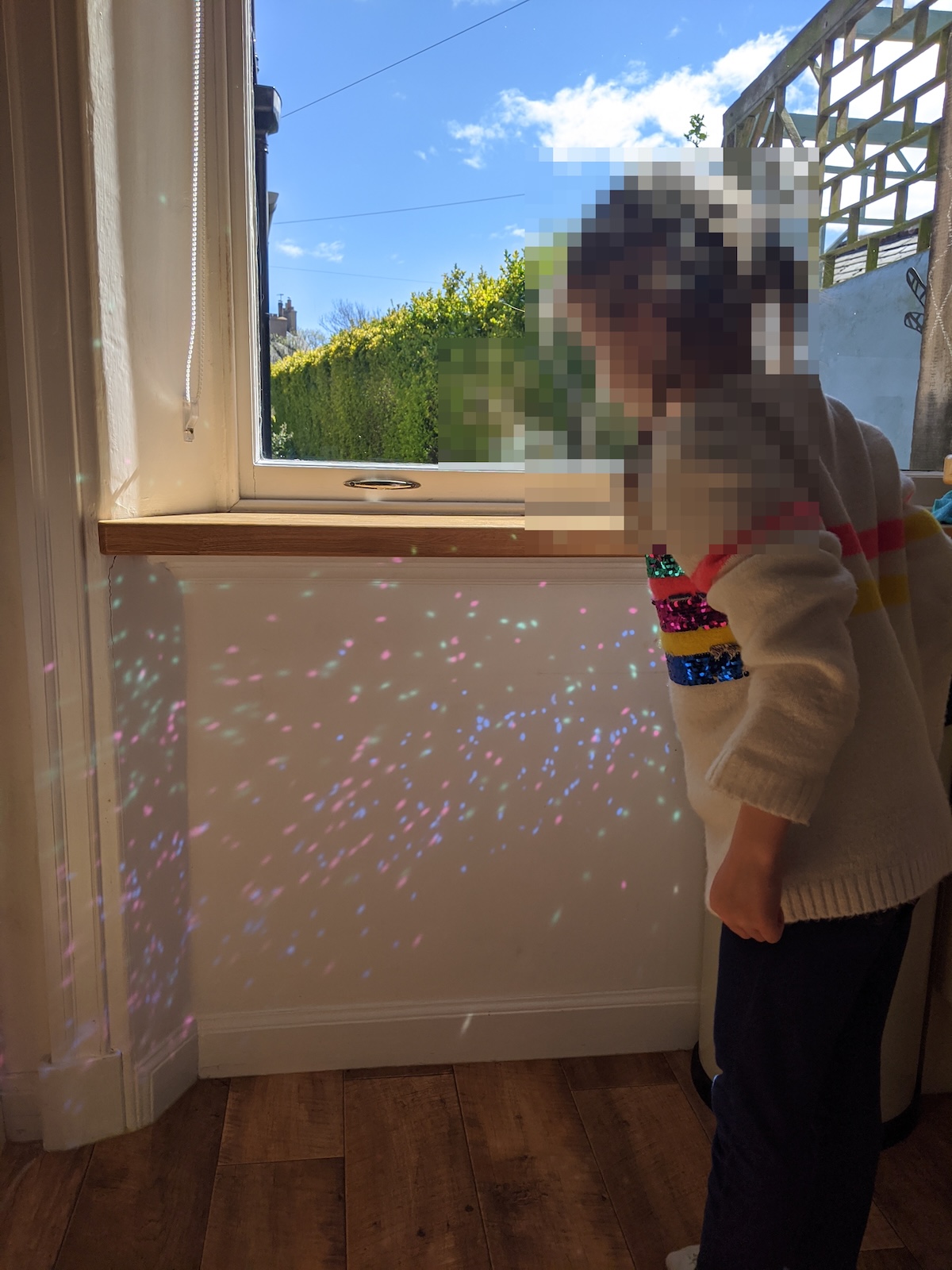 A young child standing near a sunny window as colourful rainbow light reflections speckle the wall beside them.