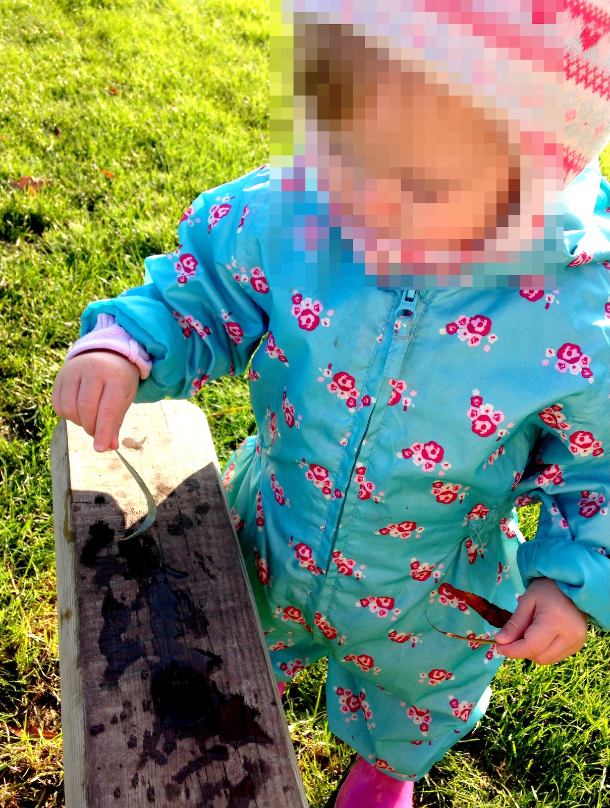 A young child wearing a blue floral puddle suit and pink patterned hat plays outdoors, touching leaves to a wooden beam over green grass.