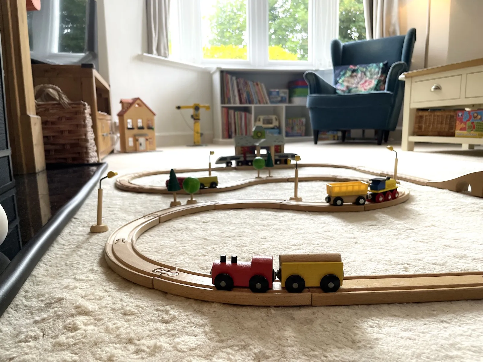 Toy trains on wooden tracks tracks in sitting room which includes other toys in the background such as books on low level shelving, a wooden, yellow toy crane and small dolls house.